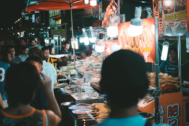 A busy night market scene with several food stalls illuminated by bright lights. Multiple people are gathered around, some observing the food selection, while others engage in transactions. The food stalls feature a variety of grilled items on skewers. Signs written in a mix of languages can be seen, advertising fried and BBQ dishes.