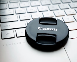 A Canon camera lens cap rests on a white HP Pavilion laptop keyboard. The keyboard is visible in the background, highlighting its keys and the laptop model name.