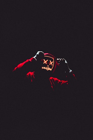 A person wearing a hoodie with a glowing mask featuring stitched and crossed-out eyes, set against a dark background. The mask emits an eerie glow, creating a sense of mystery.