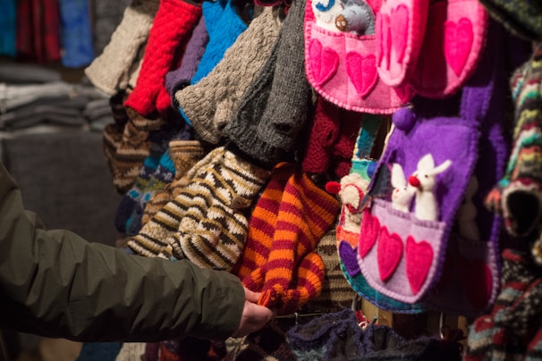 A variety of colorful knitted and crocheted items are displayed. The collection includes hats, mittens, and bags with heart and animal designs. A person’s arm is extended, touching one of the items.