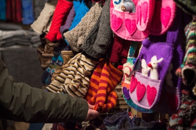 A variety of colorful knitted and crocheted items are displayed. The collection includes hats, mittens, and bags with heart and animal designs. A person’s arm is extended, touching one of the items.