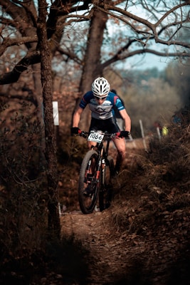 A cyclist wearing a blue and white jersey and a white helmet rides a mountain bike along a narrow dirt trail through a wooded area. The path is surrounded by trees with bare branches, indicating an autumn setting. The cyclist appears focused and is positioned in a slightly forward-leaning stance, suggesting movement and effort.