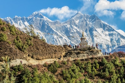 A picturesque mountain landscape features snow-capped peaks under a bright blue sky. In the foreground, a stupa with prayer flags is perched on a hillside. There are several people along a trail leading to the stupa, surrounded by green shrubs and small trees.