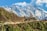 A picturesque mountain landscape features snow-capped peaks under a bright blue sky. In the foreground, a stupa with prayer flags is perched on a hillside. There are several people along a trail leading to the stupa, surrounded by green shrubs and small trees.