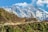 A picturesque mountain landscape features snow-capped peaks under a bright blue sky. In the foreground, a stupa with prayer flags is perched on a hillside. There are several people along a trail leading to the stupa, surrounded by green shrubs and small trees.