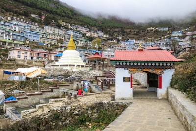 A hillside town with buildings stacked along the slope, featuring a prominent white stupa with a golden spire. A gateway with traditional architecture and colorful prayer flags add to the cultural ambiance. The landscape is lush with greenery and the sky is overcast.