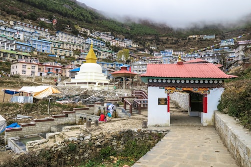 A hillside town with buildings stacked along the slope, featuring a prominent white stupa with a golden spire. A gateway with traditional architecture and colorful prayer flags add to the cultural ambiance. The landscape is lush with greenery and the sky is overcast.