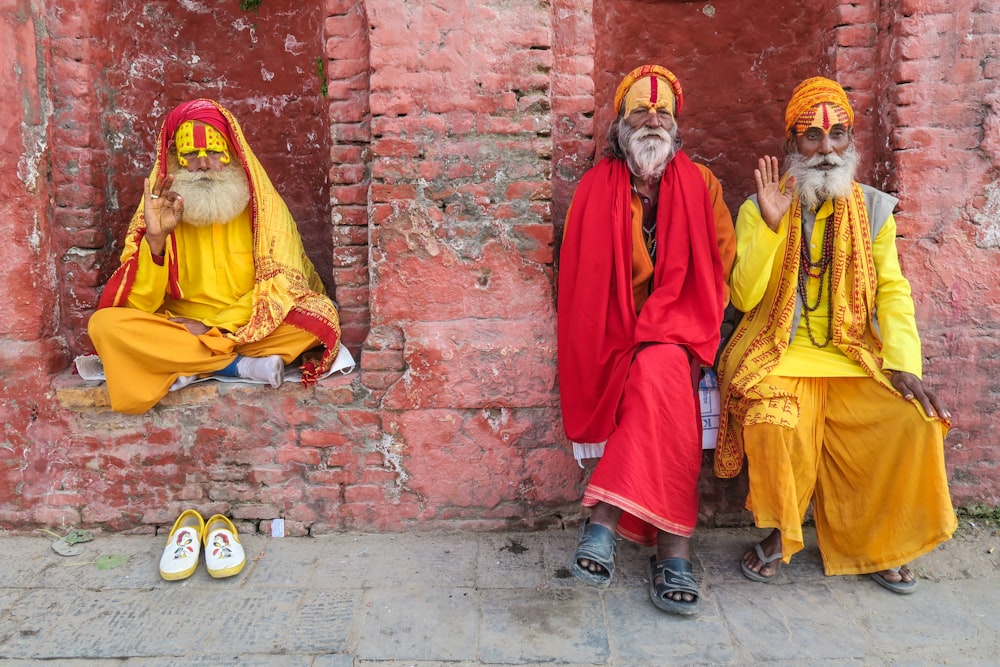 黄色と赤のローブを着た3人の宗教家が、赤い崖っぷちの建物のそばに座っている