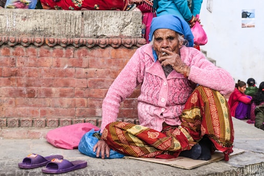 An elderly person sits against a brick wall, wearing a bright pink knit sweater, colorful patterned pants, and a blue headscarf. In front of them, pink slippers rest on the ground. Other people are seated in the background, suggesting a social or public setting.