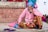 An elderly person sits against a brick wall, wearing a bright pink knit sweater, colorful patterned pants, and a blue headscarf. In front of them, pink slippers rest on the ground. Other people are seated in the background, suggesting a social or public setting.