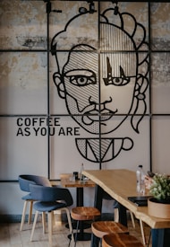 A cafe interior featuring a large mural on the wall depicting an abstract human face with geometric patterns. The words 'COFFEE AS YOU ARE' are displayed next to the mural. The room includes modern furniture with blue chairs and wooden stools around tables. A bottle and a plant are placed on a long wooden table, contributing to the cozy ambiance.
