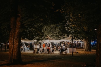 A nighttime outdoor gathering under illuminated tents, surrounded by large trees. People are seated and standing, engaging in conversations and enjoying the atmosphere.