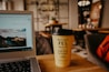 A cozy caf&eacute; setting with a close-up of a laptop showing a scenic image, next to a coffee cup with humorous text. The background features blurred furnishings and wall art, giving a warm, inviting atmosphere.