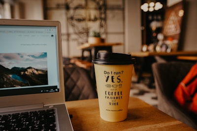 A cozy caf&eacute; setting with a close-up of a laptop showing a scenic image, next to a coffee cup with humorous text. The background features blurred furnishings and wall art, giving a warm, inviting atmosphere.