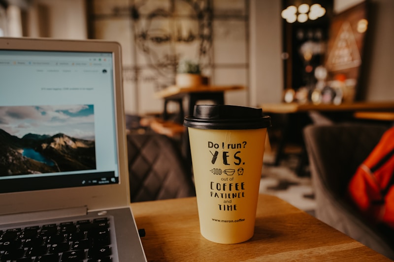 A cozy caf&eacute; setting with a close-up of a laptop showing a scenic image, next to a coffee cup with humorous text. The background features blurred furnishings and wall art, giving a warm, inviting atmosphere.