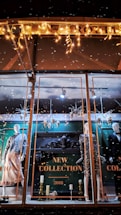 A store window display showcases mannequins dressed in stylish evening attire. The background features ornate decorations and a sign reading 'New Collection 2018.' Festive lighting and decorative elements like snowflakes and metallic ornaments add a seasonal touch. The scene is elegantly arranged with a focus on fashion and holiday ambiance.