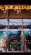 A store window display showcases mannequins dressed in stylish evening attire. The background features ornate decorations and a sign reading 'New Collection 2018.' Festive lighting and decorative elements like snowflakes and metallic ornaments add a seasonal touch. The scene is elegantly arranged with a focus on fashion and holiday ambiance.