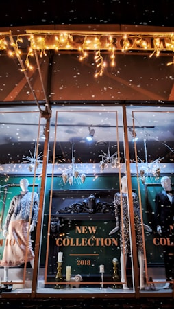 A store window display showcases mannequins dressed in stylish evening attire. The background features ornate decorations and a sign reading 'New Collection 2018.' Festive lighting and decorative elements like snowflakes and metallic ornaments add a seasonal touch. The scene is elegantly arranged with a focus on fashion and holiday ambiance.