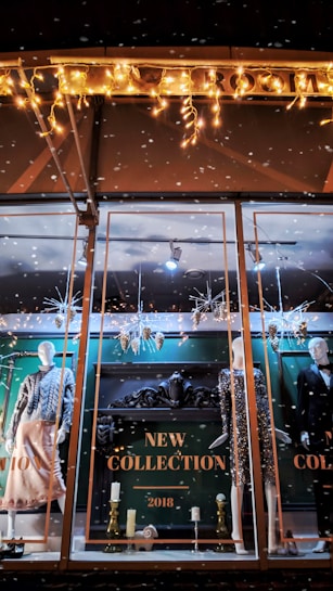 A store window display showcases mannequins dressed in stylish evening attire. The background features ornate decorations and a sign reading 'New Collection 2018.' Festive lighting and decorative elements like snowflakes and metallic ornaments add a seasonal touch. The scene is elegantly arranged with a focus on fashion and holiday ambiance.