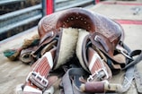 A well-worn leather saddle with intricate detailing resting on a wooden surface. The underside is padded with fur, and several straps and buckles are visible. The saddle is surrounded by other pieces of equipment, and the background shows metal railings.