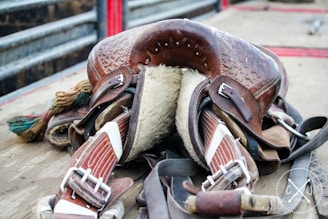 A well-worn leather saddle with intricate detailing resting on a wooden surface. The underside is padded with fur, and several straps and buckles are visible. The saddle is surrounded by other pieces of equipment, and the background shows metal railings.
