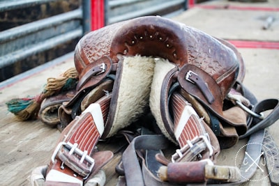 A well-worn leather saddle with intricate detailing resting on a wooden surface. The underside is padded with fur, and several straps and buckles are visible. The saddle is surrounded by other pieces of equipment, and the background shows metal railings.