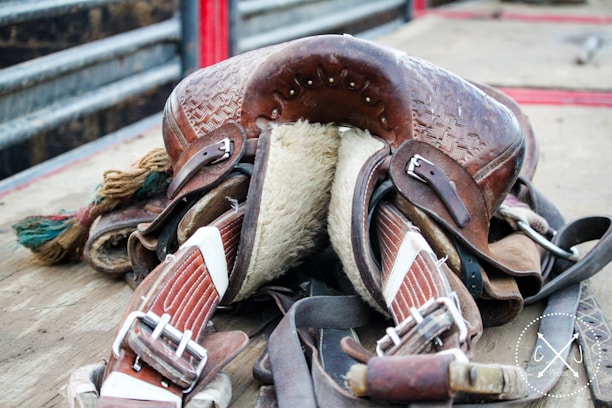 A well-worn leather saddle with intricate detailing resting on a wooden surface. The underside is padded with fur, and several straps and buckles are visible. The saddle is surrounded by other pieces of equipment, and the background shows metal railings.