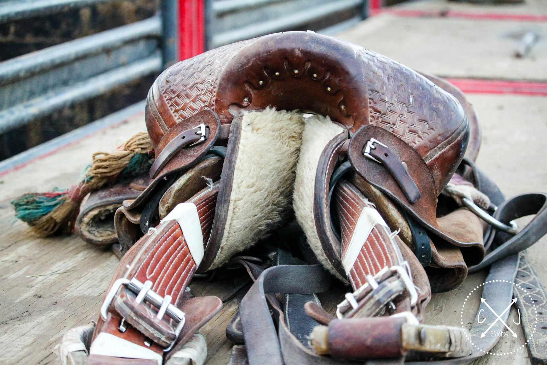A well-worn leather saddle with intricate detailing resting on a wooden surface. The underside is padded with fur, and several straps and buckles are visible. The saddle is surrounded by other pieces of equipment, and the background shows metal railings.