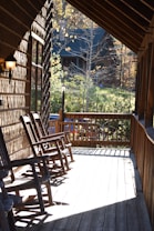 A wooden porch with a few wooden rocking chairs arranged in a row, positioned to face the surrounding natural landscape. The porch is connected to a rustic wooden house and is partially in shadow due to the roof overhang. In the background, there is another wooden house surrounded by trees and greenery, indicating a serene, wooded area. A lamp post stands at the edge of the porch, adding to the cozy atmosphere.