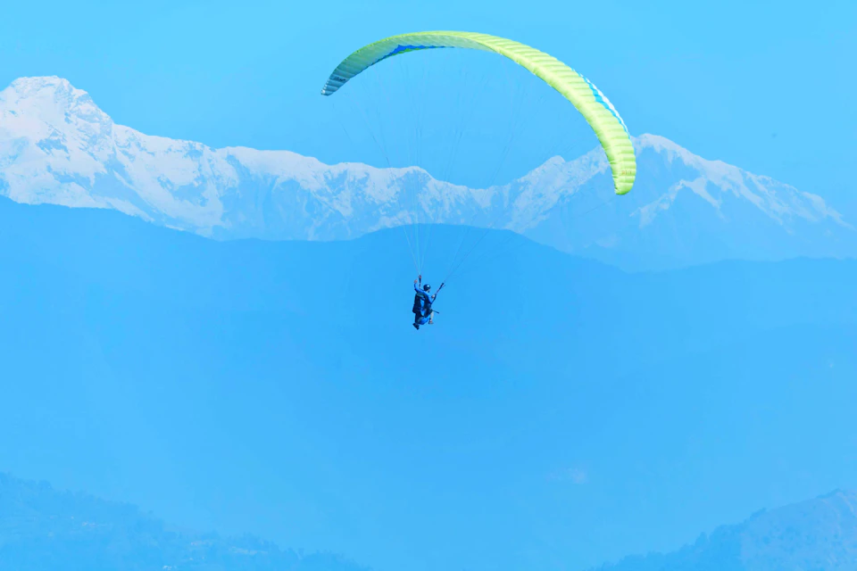 The image depicts a person paragliding over a mountain range, with their body suspended in mid-air and attached to a parachute. The paraglider is positioned centrally in the frame, facing towards the right side of the image. Below them, the majestic mountains stretch across the horizon, their peaks touching the clear blue sky.