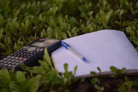 A calculator, a blank notebook, and a pen are placed on the ground amidst lush green grass. The scene captures a blend of study tools and nature.