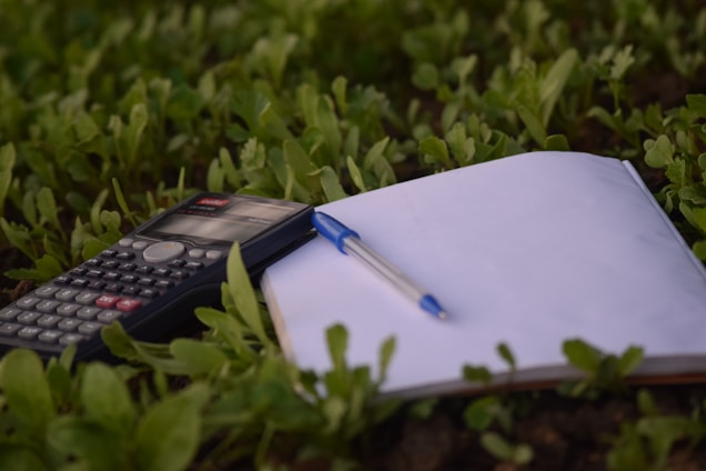 A calculator, a blank notebook, and a pen are placed on the ground amidst lush green grass. The scene captures a blend of study tools and nature.