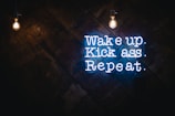 A motivational quote in neon sign format is displayed against a dark wooden background. Two bright light bulbs hang from the top, illuminating the space with a warm glow. The neon text is vibrant and eye-catching, conveying a message of determination and action.