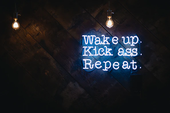 A motivational quote in neon sign format is displayed against a dark wooden background. Two bright light bulbs hang from the top, illuminating the space with a warm glow. The neon text is vibrant and eye-catching, conveying a message of determination and action.