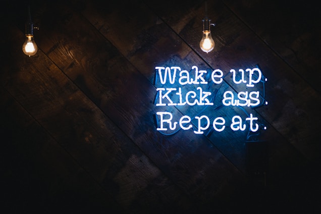 A motivational quote in neon sign format is displayed against a dark wooden background. Two bright light bulbs hang from the top, illuminating the space with a warm glow. The neon text is vibrant and eye-catching, conveying a message of determination and action.