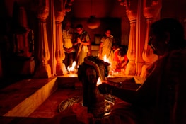 A group of people are engaged in a ritual or prayer ceremony inside a temple. The scene is illuminated by firelight, casting warm, orange glows on the ornate pillars and architecture. Some individuals are standing with hands clasped in prayer, while others are tending to the flames or the central object of focus.