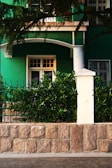 A green building facade with a white door and window is partially visible, surrounded by lush greenery. A stone wall with a white post supports decorative iron fencing, while branches overhang the top of the scene.