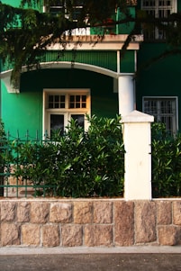 A green building facade with a white door and window is partially visible, surrounded by lush greenery. A stone wall with a white post supports decorative iron fencing, while branches overhang the top of the scene.