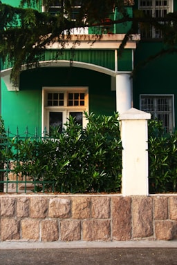 A green building facade with a white door and window is partially visible, surrounded by lush greenery. A stone wall with a white post supports decorative iron fencing, while branches overhang the top of the scene.
