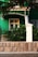 A green building facade with a white door and window is partially visible, surrounded by lush greenery. A stone wall with a white post supports decorative iron fencing, while branches overhang the top of the scene.