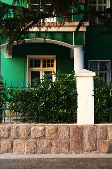 A green building facade with a white door and window is partially visible, surrounded by lush greenery. A stone wall with a white post supports decorative iron fencing, while branches overhang the top of the scene.