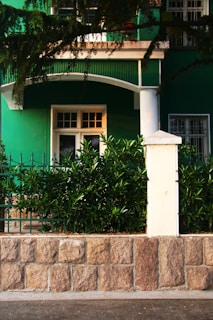 A green building facade with a white door and window is partially visible, surrounded by lush greenery. A stone wall with a white post supports decorative iron fencing, while branches overhang the top of the scene.