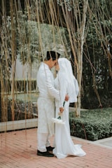 A couple dressed in traditional attire is standing closely together, sharing an intimate moment. The man is wearing a black cap and a white outfit with intricate patterns, while the woman is in a white dress and headscarf, holding a small bouquet of white flowers. They stand on a brick path surrounded by lush greenery and strings of hanging vines create a natural curtain around them.