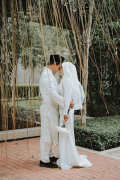 A couple dressed in traditional attire is standing closely together, sharing an intimate moment. The man is wearing a black cap and a white outfit with intricate patterns, while the woman is in a white dress and headscarf, holding a small bouquet of white flowers. They stand on a brick path surrounded by lush greenery and strings of hanging vines create a natural curtain around them.