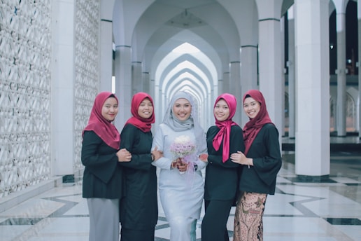 Five women are standing together in an architectural setting with white columns and arches. The woman in the center is wearing a light gray or white dress and holding a bouquet of flowers, while the others are wearing black outfits with red headscarves.