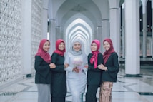 Five women are standing together in an architectural setting with white columns and arches. The woman in the center is wearing a light gray or white dress and holding a bouquet of flowers, while the others are wearing black outfits with red headscarves.