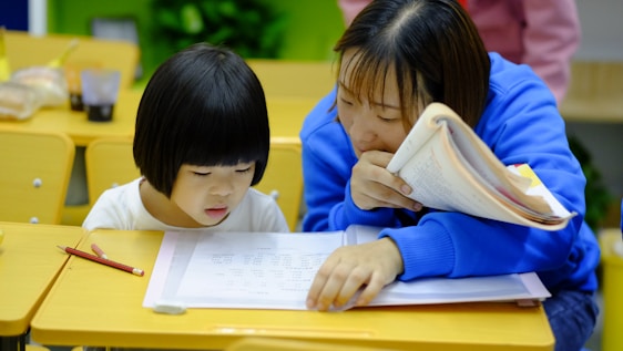 A young child and an adult are sitting at a wooden desk in a classroom, focusing on some written materials. The adult is holding a notebook and appears to be helping the child, who is intently looking at papers on the desk. Nearby are pencils and other school supplies.
