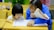 A young child and an adult are sitting at a wooden desk in a classroom, focusing on some written materials. The adult is holding a notebook and appears to be helping the child, who is intently looking at papers on the desk. Nearby are pencils and other school supplies.