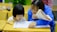 A young child and an adult are sitting at a wooden desk in a classroom, focusing on some written materials. The adult is holding a notebook and appears to be helping the child, who is intently looking at papers on the desk. Nearby are pencils and other school supplies.