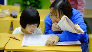 A young child and an adult are sitting at a wooden desk in a classroom, focusing on some written materials. The adult is holding a notebook and appears to be helping the child, who is intently looking at papers on the desk. Nearby are pencils and other school supplies.